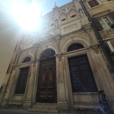 The image depicts the ornate, light-colored stone facade of the Loggia dei Mercanti (Merchants' Lodge) in Ancona, Italy. The building exhibits Venetian Gothic architectural elements, including intricate carvings, tracery, and multiple arched openings.

The central feature is a large, dark brown, double wooden door, deeply carved with decorative panels and set within an arched entrance frame. This main entrance is accessed by a small flight of three stone steps. Flanking the central doors are two tall, narrow arched windows, each fitted with dark metal grilles. Above these windows and the main door, the facade is heavily ornamented with sculptural reliefs, smaller niches containing statues (some partially obscured), and geometric patterns. Portions of an inscription, "CONSILIUM ANSARUM" and "CONSILIUM MERCATORUM", are visible on the upper facade, identifying the historical purpose of the structure. The topmost section of the building features more intricate stone work and several small pinnacles against a clear blue sky.

Direct sunlight from the upper left creates significant lens flare, partially obscuring the upper left portion of the building and the sky. To the immediate right, an adjacent building with a plainer, lighter-colored facade and closed reddish-brown window shutters is visible. An external air conditioning unit is mounted on this adjacent structure.

In the lower right corner, a single male individual is partially visible. He has dark hair, a beard, and is wearing a dark collared shirt, possibly a uniform. He appears to be walking from left to right, holding a blue object in his left hand, and is looking slightly downwards and to his right. He is partially obscured by the sun's glare. The ground in the foreground is paved, leading up to the building's steps. No other individuals or significant interactions are present.