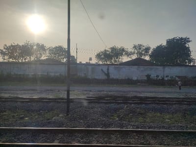 The image shows a view of a train track in Kecamatan Waru, Indonesia, with a concrete wall and trees in the background. The sun is shining brightly in the sky, and the ground is covered in gravel. A person can be seen walking on the ground near the wall, which is painted white and has some graffiti on it. There are several sets of train tracks in the foreground. The scene appears to be taken in the early morning, with a clear sky and a bright sun. The image was likely taken from a train. The environment is serene and quiet, with no other people or vehicles visible.