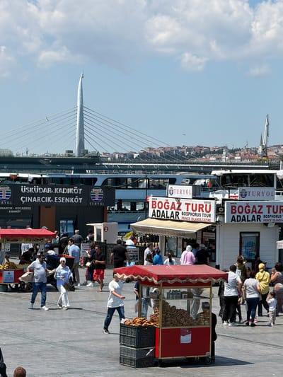 The image depicts a busy outdoor scene in the Eminönü district of Fatih, Turkey, on a sunny day with scattered clouds. In the background, a large cable-stayed bridge spans the Golden Horn, with a white ferryboat docked in the foreground, displaying "ÜSKÜDAR-KADIKÖY ÇINARCIK-ESENKÖY" and "TURYOL" on its side. Several people are visible on the ferry and on the pier.
Closer to the foreground, multiple food carts are set up. One cart is red with a fringed awning and is filled with simit (Turkish bagels). Another cart is white with red signage advertising "BOĞAZ TURU ADALAR BOSPHORUS TOUR PRINCE ISLANDS SEZİRE." Several people are gathered around these carts, some appearing to be customers and others vendors.
People of various ages are walking around the paved area, dressed in casual summer clothing. Some are carrying bags or purchasing items from the carts. The overall atmosphere is one of a bustling tourist area with ongoing activity.