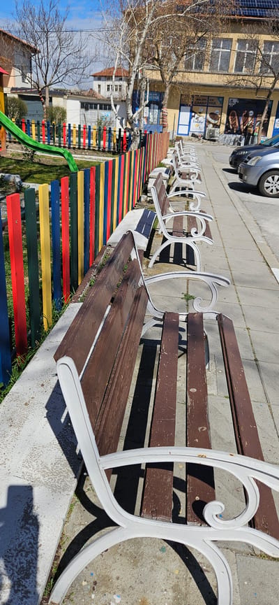 The image shows a row of park benches located in Slatino, Bulgaria, situated along a walkway next to a playground. The benches are made of brown wooden planks with white metal frames. The playground features a colorful fence with horizontal stripes of red, yellow, blue, green, and red. A green slide is visible on the left side of the image. Beyond the playground, there are buildings, bare trees, and a car parked on the street. The image is taken from a slightly elevated angle, capturing the benches and playground in the foreground.