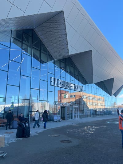 The image depicts the exterior of a modern airport terminal building under a clear blue sky in Murmashi, Russia. The building features an angular design with large reflective glass panels on the lower facade and light-gray metallic panels, some perforated, on the upper structure. A prominent sign on the glass reads "НА СЕВЕРЕ ЖИТЬ" in large white capital letters, with "ВХОД ENTRANCE" and an arrow below it, indicating the main entry.
In the foreground, a dark gray paved area shows two circular manhole covers. Several individuals are present:
* On the far left, a metal luggage cart is visible.
* Near the left side of the building, a wooden bench holds two seated individuals; one wears a dark jacket and beanie, the other dark clothing. Two dark wheeled suitcases rest on the pavement in front of them.
* A third individual, wearing a dark jacket and beanie, stands near the bench.
* Two females walk towards the right; one in a light jacket carries a dark shoulder bag, the other in a dark jacket has a pink item draped over her shoulder.
* Multiple silver luggage carts are parked near the glass entrance doors.
* On the far right, a person wearing an orange and black jacket is partially visible, cropped at the waist.
The glass facade reflects internal lighting, the blue sky, and an older, multi-story light-brown brick building located opposite the terminal.