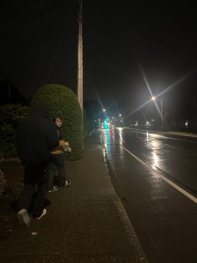 The image displays an outdoor nighttime scene in Saanich, Canada, under wet conditions, suggesting recent or ongoing rain. Two individuals are positioned on a grey concrete sidewalk adjacent to a wet asphalt road.
Subject 1, in the left foreground, is partially visible from the mid-thigh down, wearing dark pants and light-colored sneakers, appearing to be in motion towards the right. The upper body is obscured by dark clothing or shadow.
Subject 2 stands to the right of Subject 1, facing slightly towards the camera. This individual wears a yellow/tan jacket over dark pants and dark sneakers with white soles. A green bottle is held in their left hand. This subject appears to be a young male with brown hair.
Along the left side of the sidewalk, a dense, manicured green hedge forms a barrier. Behind the hedge, a wooden utility pole with wires is visible. The wet asphalt road, marked with a solid white line, reflects multiple streetlights and distant traffic lights displaying green and red hues. A prominent streetlight with a starburst flare is visible on the right side of the road in the mid-distance. Further down the road, a white rectangular sign with a red circle and a diagonal line (prohibitory symbol) is discernible on the right shoulder.
