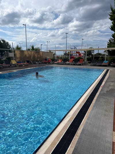 In Ayvalık, Turkey, a person is swimming in a bright blue swimming pool on a cloudy day. The pool is surrounded by a paved deck with lounge chairs and umbrellas. In the background, a playground with slides is visible. The scene appears to be at a resort or hotel.
