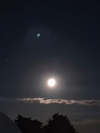 The image depicts a night sky above Passignano sul Trasimeno, Italy. A bright, full moon dominates the center of the frame, its light illuminating a thin band of clouds below. Silhouetted trees are visible in the foreground, along with a portion of a white structure on the left. Several celestial bodies, appearing as small, bright dots, are scattered across the dark sky, with one larger, pale green celestial body situated in the upper left quadrant. The overall scene suggests a clear night with a prominent moon.