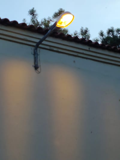 A street lamp illuminates a light-colored wall at dusk in Nea Raedestos, Greece. The lamp, affixed to the building's exterior, is angled downwards, casting a warm yellow glow that creates two large, soft shadows on the wall below. The building has a dark-tiled roof with red clay tiles, and a white facade with horizontal decorative lines. Above the roof, the silhouettes of evergreen trees are visible against a pale blue sky. The overall atmosphere is calm and tranquil, suggesting the transition from day to night. There is no visible text in the image.