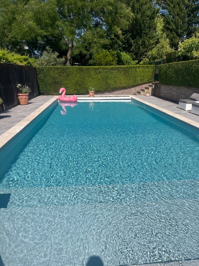 A swimming pool is filled with clear blue water, reflecting sunlight. A pink inflatable flamingo floats on the surface of the water. The pool is surrounded by a paved patio. In the background, there is a neatly trimmed green hedge and several trees. A brick wall is visible behind the hedge. There are two planters with white flowers, one on the left side of the pool and another on the right side near the flamingo. Further to the right, there are two white outdoor chairs with grey cushions. The scene appears to be a private backyard in Saint-Nom-la-Bretèche, France, on a sunny day.