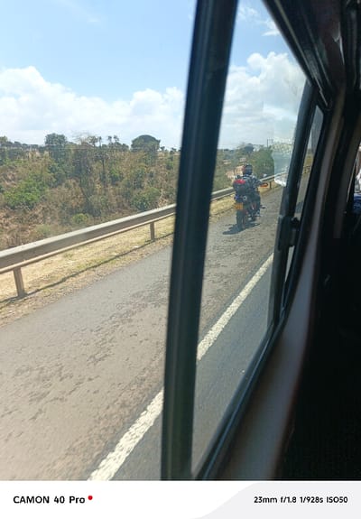 The image depicts an outdoor roadside scene in Gilgil ward, Kenya, captured from within a moving vehicle through a partially open window. A black asphalt road with a solid white edge line extends into the mid-ground. Parallel to the road's left edge, a silver metal guardrail with vertical support posts is visible. Beyond the guardrail, the terrain slopes downward, covered with sparse dry vegetation, shrubs, and trees under a bright blue sky with scattered white cumulus clouds. A single individual, wearing a dark helmet and clothing, is operating a red and black motorcycle equipped with a rear luggage box, proceeding away from the camera in the right lane. The motorcycle bears a yellow Kenyan license plate displaying "KXY" followed by numeric characters. The sunlight is bright, creating visible shadows.