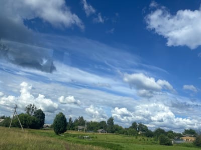 The image depicts a rural landscape under a dynamic sky with both wispy cirrus clouds and larger cumulus formations against a vibrant blue backdrop. The foreground is dominated by a green field with tall grasses, leading to a gentle slope. In the middle ground, a scattered collection of trees and vegetation borders a settlement. Several small, rustic buildings are visible, some appearing to be wooden houses with dark roofs, and one structure is in a state of construction with exposed wooden beams. A utility pole with wires stands prominently on the left side of the field, with its wooden support structure clearly visible. The overall impression is that of a tranquil, rural setting in the vicinity of Грозаўскі сельскі Савет, Belarus, likely during daylight hours with pleasant weather.