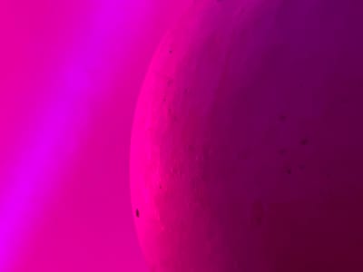 The scene, set within the city of Derby, United Kingdom, presents a close-up, abstract view dominated by shades of magenta and pink. A large, curved object, appearing spherical or ovoid, occupies the right portion of the frame. Its surface features an irregular, textured topography with multiple small, dark, circular indentations or blemishes scattered across it. This object is primarily illuminated from the upper-left, creating a vibrant magenta hue on its left curve that gradually deepens into a rich, dark magenta towards its right. The background, occupying the left side of the image, consists of a solid field of bright magenta light, smoothly blending with the tones of the foreground object. The entire visible environment is saturated in these intense pink and magenta colors.