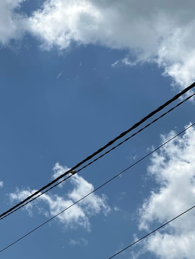 The image shows a view looking upwards towards a bright blue sky with scattered white clouds. Several black power lines diagonally traverse the frame from the upper left to the lower right. A bird is perched on one of the power lines. The location is implied to be Aprilovo, Bulgaria, but this detail is not visually verifiable within the image. No other people, subjects, objects, actions, or interactions are visible.
