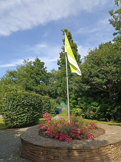 A bright green flag with white lettering is planted on a flagpole in front of a circular stone planter filled with pink flowers and green foliage. The flag is positioned in the center of the image, angled towards the right. Behind the flag and planter, a dense backdrop of lush green trees and bushes creates a natural setting. The sky above is a clear blue with scattered white clouds. The scene appears to be outdoors, likely in a park or garden area. The flag has the word "Community" printed on it in white text, and a graphic of what appears to be a stylized leaf or hand. The stone planter is constructed from irregularly shaped, flat stones stacked in a circular fashion. The surrounding ground is covered with gravel. The environment suggests a well-maintained outdoor space, possibly a public area within the town of Dungloe, Ireland, given the context.