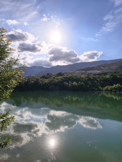 The image depicts a serene natural landscape with a focus on a calm body of green water reflecting the sky and surrounding environment. In the foreground, on the left, is a lush green tree branch with leaves, partially obscuring the view. The main subject is a wide expanse of water, its surface smooth and glassy, mirroring the bright blue sky dotted with fluffy white and gray clouds. The sun shines brightly, creating a strong glare on the water's surface and casting a luminous glow in the sky.

Behind the water, a dense, verdant forest covers the slopes of rolling hills, which in turn are backed by the soft, hazy outlines of distant mountains. The forest appears thick and vibrant with a multitude of trees. The reflection in the water clearly shows the shapes of the clouds and the sun, giving the scene a sense of depth and tranquility.

The time of day appears to be late morning or early afternoon, given the position and intensity of the sun. The weather seems fair with scattered clouds, contributing to the picturesque view. The overall mood of the image is peaceful and idyllic, emphasizing the beauty of nature. There are no visible people or man-made objects apart from what might be a very small, indistinct shape on the far right of the water's surface, possibly a small boat or debris, and a hint of a road or clearing on the hillside. There is no visible text in the image.