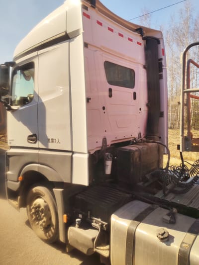 A white semi-truck (tractor unit) is parked outdoors on what appears to be an asphalt surface under a clear blue sky. The image captures the side and rear of the truck's cab and chassis, illuminated by bright daylight, casting distinct shadows. The background features several bare deciduous trees, suggesting a late autumn, winter, or early spring season in a rural or natural setting.

The truck's cab is predominantly white, with a sleeper compartment visible at the rear, featuring a small window. Red reflective strips are applied horizontally across the back of the cab. A black exhaust stack runs vertically up the rear of the cab. Below the cab, parts of the chassis are visible, including a dark grey fuel tank, a white air filter canister with Cyrillic text, and coiled air or electrical lines. The front wheel and mudguard are also visible.

No people are present, and the truck appears stationary, with no specific activity taking place.

Visible text on the side of the cab includes "HOWO T7H" faintly printed, and more clearly, "准乘2人" (Zhǔn chéng 2 rén), which translates to "Authorized to carry 2 people" or "Capacity 2 persons". On the white canister, partially visible Cyrillic text reads "ЭЛЕМЕНТ ФИЛЬТРУЮЩИЙ" (Element Filtering).

The scene is set in Novoe Mart'yanovo, Russia.