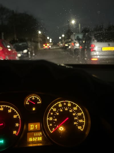 The image depicts a nighttime scene observed from the interior of a stationary vehicle, located on a wet street in Gillingham, United Kingdom. Raindrops are visible on the vehicle's windshield, obscuring the view.
The foreground displays the illuminated dashboard instruments of the vehicle. The speedometer, ranging from 0 to 160 mph (with an inner scale of 0 to 240 km/h), shows the needle resting at 0, indicating the vehicle is not in motion. A yellow "check engine" warning light is illuminated on the speedometer face. To the left, a tachometer is partially visible, also at 0 RPM. Above the tachometer, a fuel gauge indicates the fuel level is slightly above empty. A central digital display shows "D1" (likely current gear), "1117" (potentially time or trip duration), and an odometer reading of "163238". A green light is illuminated on the far left of the dashboard.
Through the rain-speckled windshield, a blurred urban or suburban street is visible. Multiple vehicles are present, indicated by their illuminated headlights and taillights. A red vehicle is partially visible on the left, and a white van with a yellow UK license plate is on the right, displaying illuminated brake lights. Streetlights provide ambient illumination along the road, casting reflections on the wet surface. No individuals are discernible in the scene.