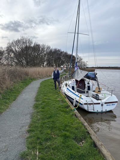 An outdoor scene captured on an overcast day depicts a male individual standing beside a moored white sailing yacht on a calm waterway, likely located within the Norfolk Broads in the district of North Norfolk, United Kingdom. The man, with dark hair and a beard, is positioned on a narrow strip of green grass separating a gravel path from the water. He wears a dark sleeveless puffer vest over a light-colored long-sleeved shirt and dark trousers, and is facing the camera with a subtle smile, his right hand slightly raised in a gesture. The sailboat, displaying the name "Misty Blue" on its transom and "Regal 525" on its hull, features a single mast, an outboard motor mounted on the stern, and a grey tarp covering a portion of its cabin area. Yellow mooring lines connect the boat to the wooden embankment. A light grey gravel path curves gently to the left alongside the grassy bank. The background consists of tall, dry, light-brown reeds and a dense line of leafless deciduous trees. Distant reed beds and a faint structural silhouette are visible on the far right horizon under a cloudy sky. The water is brownish and still.