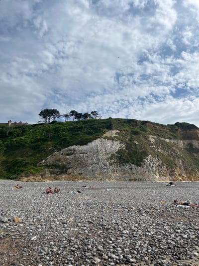 The image depicts a pebble beach at Hautot-sur-Mer, France. The scene is dominated by a steep, grassy cliff face on the left, with a white, rocky section visible partway up. Several people are scattered across the beach, appearing to be sunbathing or relaxing. Some are lying on towels, while others are seated.

The sky is a light blue with scattered white clouds, suggesting it is daytime, likely late morning or early afternoon. The weather appears fair. The vast expanse of pebbles forms the foreground and covers most of the beach area. A few trees are sparsely growing along the top of the cliff. In the distance, a small structure, possibly a building or part of a path, is partially visible at the base of the cliff. There is no visible text in the image. The overall impression is one of a peaceful, leisurely day at the seaside.