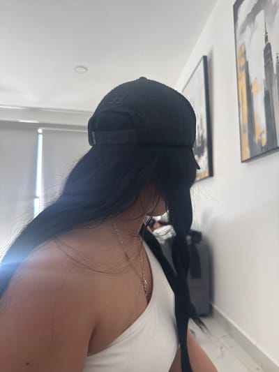 A medium shot shows a person wearing a black baseball cap and a white tank top, with long black hair in a braid. A thin, silver necklace is visible around the neck. Two framed paintings hang on the wall, one with gray and gold strokes, and the other in grayscale. A white wall with a window and a gray object in the background complete the scene. The location is inside an unidentified building in Tequesquitengo, Mexico.