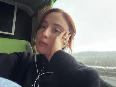 A female subject with reddish-brown hair is depicted in a medium-shot, seated inside a vehicle. The location is stated as Moineville, France, though no specific geographical identifiers are visible in the image. The subject is looking directly forward. Her right hand, adorned with two silver-colored rings on the ring and middle fingers, is positioned against her cheek. She wears a dark, long-sleeved top featuring multiple metallic studs down the front. White wired earbuds are present, with one earpiece in her left ear. To her left, a portion of a green and grey upholstered seat is visible, along with a dark wall housing a white-framed rectangular screen. The right side of the frame features a window displaying an overcast exterior. Rain or condensation streaks are visible on the windowpane, partially obscuring a distant, hilly landscape under a bright, diffused sky. The interior lighting is subdued.