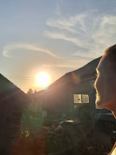 The image shows a low-angle view, looking up. In the foreground is the partial view of a person's face. The person is looking toward a bright sun that is partially obscured by a structure resembling a building. There are also some white clouds in the sky. The area around the building looks like a cluttered area. Based on the specified location, this appears to be a snapshot in or near Шешурга, Russia.