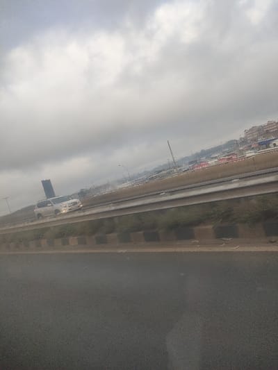 This image depicts an urban or suburban multi-lane highway scene in Gatongora ward, Kenya, under heavily overcast conditions. The perspective is from a moving vehicle, looking forward and slightly to the right across multiple roadways.
The sky occupies the upper two-fifths of the frame, filled with dense, light grey to white clouds, indicating an overcast day. Diffused ambient light illuminates the scene.
In the immediate foreground, the surface of a dark asphalt road is visible, appearing dark and slightly reflective, possibly wet. This road transitions to a raised median or divider constructed of concrete, painted with alternating dark and light (possibly black and cream/white) rectangular segments. Behind this curb, a strip of sparse, low-lying green and brown vegetation grows on an earthen slope.
Above this slope, an elevated asphalt roadway runs parallel, equipped with a metal guardrail on its side closest to the viewer. A light-colored SUV, possibly silver or white and resembling a Toyota Land Cruiser Prado, is in motion on this elevated roadway, traveling from left to right. Its headlights are illuminated. A dark, tall, rectangular object is visibly secured to the vehicle's roof rack or rear, extending vertically. The surface of this elevated road also exhibits reflective properties, suggesting dampness.
In the background, beyond the elevated roadway, various structures are visible. To the far left, a tall, dark-colored multi-story building with a distinctive architectural profile stands in the distance. Further to the right, a cluster of commercial or residential buildings is present, with several prominent red and white signs, one partially legible as "MAL..." on a red background. Utility poles, streetlights, and a distant crane-like structure or communication tower are also discernible. No individuals are visible in the frame.