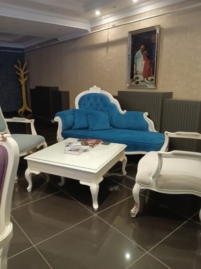 The image depicts a luxurious waiting area, possibly in a hotel or upscale establishment, in the city of Kırıkkale, Turkey. The scene is dominated by ornate furniture, including a vibrant blue chaise lounge with tufted upholstery and white decorative trim, flanked by two white armchairs with similar detailing. A white, glass-topped coffee table sits in the center of the arrangement, holding a stack of books. The floor is covered in dark, glossy tiles laid in a grid pattern. In the background, a framed painting hangs on a textured beige wall, and a unique, yellow tree-shaped coat rack stands against a darker wall. The ceiling features intricate molding and recessed lighting. The overall impression is one of elegance and comfort.