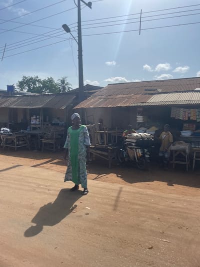 The scene depicts a street in Ikoga, Nigeria. The street is unpaved and dusty. Along one side of the street is a row of corrugated metal-roofed structures housing small shops. One shop has bags and goods stacked on a motorcycle. Several people are visible, including one person wearing a green and white patterned dress and head covering walking in the foreground. Power lines and a utility pole are visible on the left side of the frame. The sky is light blue with scattered clouds.
