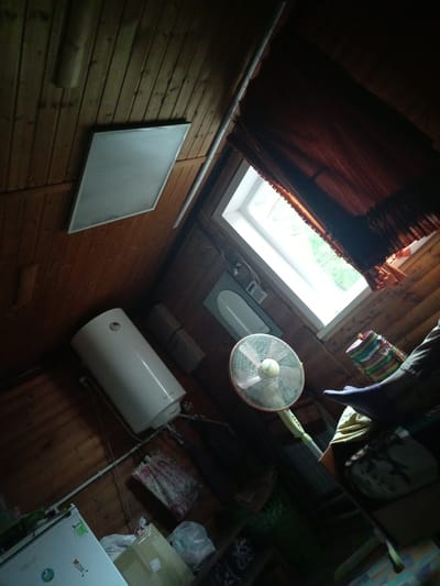 The image depicts an interior scene, likely a room with a sloped wooden ceiling and walls. In the upper left, a rectangular fluorescent light fixture is mounted on the ceiling. Below this, a large, white, cylindrical water heater is positioned vertically against the wooden wall. To the right of the water heater, there are two rectangular electrical boxes.
A significant feature of the room is a skylight window, partially obscured by dark reddish-brown curtains with fringe at the bottom. Through the window, greenery and daylight are visible. In front of the window and slightly to the left, a white oscillating fan with a pink grille is set up.
In the lower right of the frame, a person's legs and feet are visible, wearing dark pants and a purple shoe or slipper. The person appears to be resting or sitting. Next to the fan, a colorful woven cylindrical object, possibly a basket, is placed on a surface.
The floor is made of wooden planks. Various items are scattered around the room, including what appears to be a white refrigerator in the lower left corner, some boxes, and bags. The overall lighting is dim, with light emanating primarily from the skylight. The setting suggests a living space, perhaps an attic room or a cottage, in Засопка, Russia.