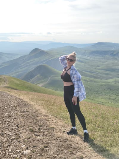 A woman stands on a dirt path in Коктебельский поселковый совет, Ukraine. She is wearing a black sports bra, black leggings, a blue and white plaid shirt, and black sneakers. She has sunglasses on and her blonde hair is in a bun. Her left hand is on the back of her head. The background consists of rolling green hills and mountains.