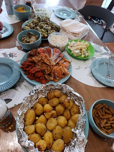 A close-up, overhead shot shows a wooden table laden with various dishes, suggesting a meal or gathering. In the foreground, a foil-lined tray overflows with golden-brown roasted potatoes seasoned with herbs. To the right, a blue bowl contains an assortment of toasted breadsticks or croutons.
Further back on the table, a blue plate displays a colorful arrangement of sliced salmon, sun-dried tomatoes, and what appears to be fried onions or crispy noodles. Adjacent to this, a green plate holds cubed cheese, some with visible blue veins, indicating a cheese selection. There are also bowls containing shredded cabbage salad and what looks like pickled mushrooms. Several blue plates with decorative patterns are scattered across the table, one with a fork resting on it. A stack of flatbread or tortillas is also visible, as is a glass bottle with a label partially obscured. The scene is lit by ambient light, possibly from a nearby window, creating a warm and inviting atmosphere. The overall impression is one of a shared meal, likely a casual gathering with friends or family. There is no discernible time of day or weather information.