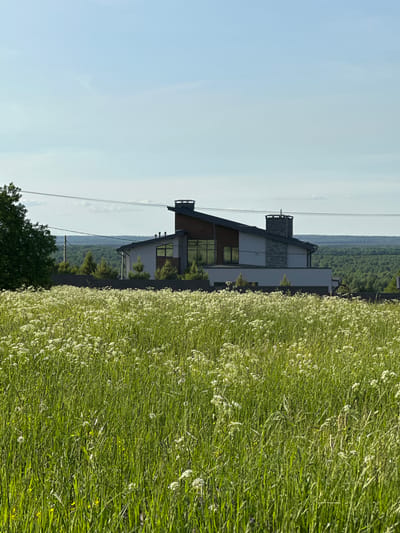 The image depicts a modern house situated in Novaya Ropsha, Russia. The house is a two-story structure with a unique, angular roof design and large, multi-pane windows. It features a stone-clad chimney. In the foreground, a vast field of tall green grass with small white flowers stretches towards the house. A dark fence separates the field from the property. Behind the house, a dense forest forms the backdrop, and power lines are visible in the sky. The sky is a clear blue with a few wispy clouds. No people are visible in the image.