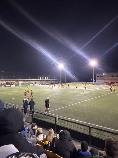 An outdoor nighttime football match is depicted at Arbour Park, Slough, United Kingdom. The image is captured from an elevated position within a spectator stand, overlooking an artificial turf pitch illuminated by multiple floodlights, which create prominent lens flares against the dark sky. Two teams of players are present on the pitch: one team wears yellow and black kits, with approximately 10-12 players primarily positioned in the mid-ground and right side of the field. The opposing team wears maroon and dark kits, with approximately 6-8 players visible on the left side of the pitch, near the penalty area. A referee in a black uniform and holding a yellow flag stands near the foreground sideline, observing the field. A football is visible on the pitch near the left penalty area. In the foreground, multiple spectators are seated in tiered wooden-style seating, separated from the pitch by a black metal barrier. They are dressed in winter clothing, including hooded jackets. Advertising banners for "SLOUGH", "VESTEL", and "HUNGER & CRAY BREWERY" are visible along the perimeter of the pitch behind the left goal. A multi-story building is visible in the background to the right.