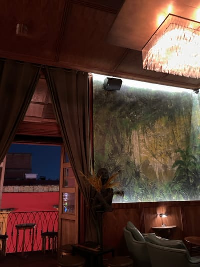 The image shows the interior of a bar or restaurant in Oaxaca de Juárez, Mexico. The bar is dimly lit, with a large mural depicting a lush jungle scene on the wall. A large, ornate chandelier hangs from the ceiling. There are several wooden tables and chairs scattered throughout the space. Two bar stools can be seen in the foreground, along with a partially obscured wooden table. A large, ornate mirror hangs on the wall near the bar, reflecting a view of the street outside. Through the window, a red-painted balcony can be seen. There is a statue of a human figure in a seated position and a small, potted plant in the corner of the room. The image appears to have been taken at night, as the sky outside is dark. The image is taken from a low angle and shows the interior of the establishment.