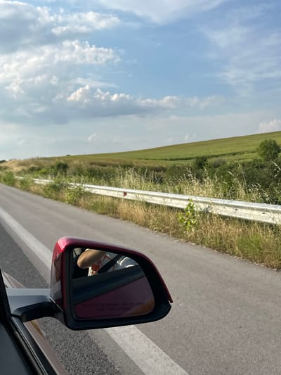 The image depicts a daylight scene observed from the front passenger side of a moving vehicle in the vicinity of Kapitan Andreevo, Bulgaria. The primary view extends along an asphalt road with a solid white line marking its right edge. To the right of the road, a white metal guardrail is present, partially obscured by an abundance of dry, yellowish grass, weeds, and green shrubbery growing roadside and behind it. Beyond the guardrail and immediate vegetation, the landscape features an expansive, upward-sloping green field or hill, indicating a rural or semi-rural environment. The distant horizon is characterized by the contours of these fields, with a few small trees visible. The sky is bright blue with scattered white cumulus clouds, suggesting fair weather.
In the foreground, occupying the lower-left quadrant, is the exterior side mirror of the vehicle. The mirror's casing is a dark red or maroon, mounted on a black base attached to the vehicle's dark-colored door frame, which is partially visible. The mirror's reflection shows the right arm and shoulder of an individual, presumably the driver, wearing a light-colored t-shirt with indistinct orange or yellow patterns. A dark seatbelt is diagonally visible across the person's shoulder and chest. The reflection also reveals part of the vehicle's interior, appearing dark red, likely a dashboard or door panel. Text is visible on the lower surface of the mirror, stating "OBJECTS IN MIRROR ARE CLOSER THAN THEY APPEAR." The texture of the asphalt road surface is visible in the lower right and foreground, appearing coarse and grey.