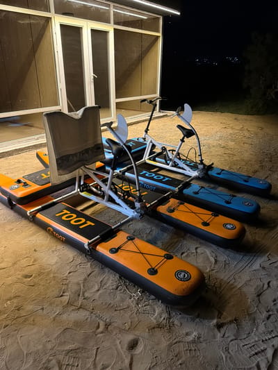 The image shows a fleet of four pedal-powered watercrafts parked on a sandy area outside a building with glass doors and windows. The watercrafts are a combination of orange and blue, with black accents and a white frame. Each watercraft has a seat, handlebars, pedals, and a propeller at the rear. The word "TOOT" is visible on the orange watercrafts. The building appears to be a rental facility or a place of business. In the background, a dark night sky is visible with distant lights, suggesting the location is a town or city. The sandy ground is covered with numerous footprints, indicating a high volume of foot traffic. The overall scene suggests a recreational area, possibly near a lake or beach, in the city of Cholpon-Ata, Kyrgyzstan.