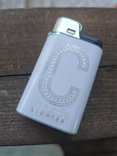 A white lighter with a decorative crystal "C" on its side and the word "LIGHTER" printed below it rests on a weathered wooden surface. The lighter is angled towards the viewer, with its chrome top and black ignition mechanism visible. The wooden surface appears to be outdoors, possibly a table or bench, with visible grain and imperfections. The background is blurred, suggesting an outdoor environment. The image was taken in Mäntsälä, Finland.