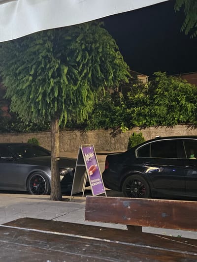 An outdoor scene at night, possibly in the city of Byala, Bulgaria, captures a street-level view. In the foreground, a dark wooden bench is partially visible, suggesting a seating area. Behind it, a street or sidewalk runs parallel to a row of buildings. The middle ground features two parked cars: a dark gray sedan on the left and a black sedan on the right. Between the cars and partially obscuring the gray sedan is a young tree with lush green foliage and a rough, textured trunk. To the right of the tree stands a purple A-frame sign advertising "Milka" chocolate, featuring an image of chocolate bars. The background consists of a stone wall and dense green bushes, with a dark, night sky above. The lighting appears to be artificial, illuminating the scene and casting shadows.