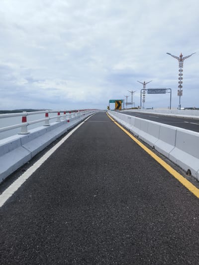 The image depicts a perspective view of a modern highway bridge under a cloudy sky. The foreground is dominated by the asphalt road surface, marked with a white line on the left and a yellow line on the right. Concrete barriers with white railings line both sides of the road. In the distance, ornate streetlights with intricate gold detailing are visible. A blue sign with white lettering and a yellow chevron sign indicating a curve are also present. The background shows a body of water and distant landmass. The scene is devoid of people or vehicles. This bridge is likely part of the Mandara toll road, connecting Benoa Bay to the Ngurah Rai Airport and the mainland of Bali, located in South Denpasar, Indonesia.