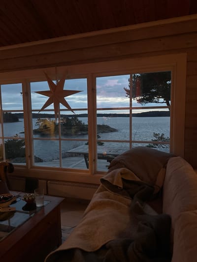 The image presents an indoor scene looking out through a multi-paned window towards a large body of water in Boda, Sweden, during twilight. The interior space features light wooden paneling on the walls and ceiling, and a light-colored wooden floor.

In the foreground, a dark wooden coffee table with a glass top is visible on the left, holding a clear glass of water, a dark bowl containing small, dark spherical items, another small bowl with silver-colored items, and folded paper. To the right, a light-colored upholstered sofa is partially visible, covered by a beige blanket and a darker throw, with a grey pillow positioned on it. Below the window, a white horizontal radiator is installed.

The window, framed in white, features a suspended eight-pointed paper star decoration in its upper-left quadrant. Beyond the window, a wide expanse of water appears calm under a sky with pale blue and grey clouds. Several rocky islets and shorelines covered with coniferous trees are visible across the water. A wooden table is present on an outdoor structure, likely a deck or dock, directly adjacent to the water beneath the window. No individuals are depicted in the scene.