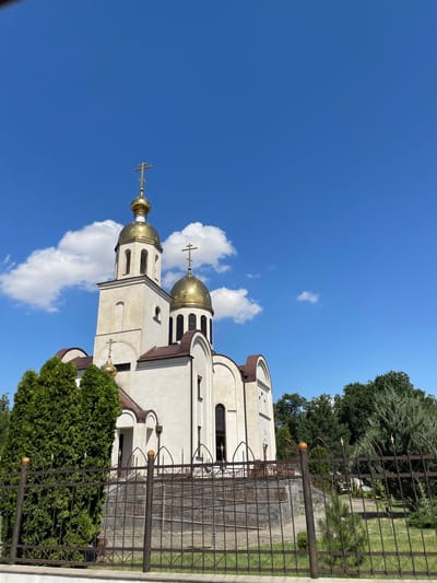The image shows a white Orthodox church with golden domes under a clear blue sky with scattered white clouds. The church has a tall bell tower and a smaller dome, both topped with crosses. A dark metal fence is in the foreground, partially obscuring a pathway leading up to the church entrance. Green trees, including a tall evergreen on the left, are visible behind the fence and around the church. The scene is set in the city of Прасковея, Russia. There are no people visible in the image.
