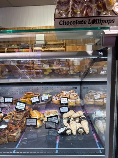 The image depicts the interior of a bakery display case located in Wyre Forest, United Kingdom, presenting a selection of baked goods for sale. The foreground features a refrigerated glass display, illuminated by an internal LED light strip, containing multiple trays of pastries and desserts. Specific items visible include square portions of "Bread Pudding" and "Quality Crust Bread Pudding" priced at £1.99 each, round "Medium Egg Custard" tarts at £2.25, "Pineapple Tart" at £1.70, and stacked "Bakewell Tart" at £3.99 each. Also present are "Apple Doughnut" at £1.99, "Synthetic Cream Strawberry Devon" at £2.25, "Synthetic Cream Apple Turnover," and a cluster of "Butter Cream Horns" at £1.99, some with chocolate-dipped ends. Above the main display, a cardboard box holds individually wrapped "Chocolate Lollipops," advertised as "Finest Belgian Chocolates." The background, visible through the display glass, shows wooden shelving units stocked with additional baked items, including bread rolls and packaged pastries, against a white paneled wall. No individuals or specific actions are depicted within the frame.
