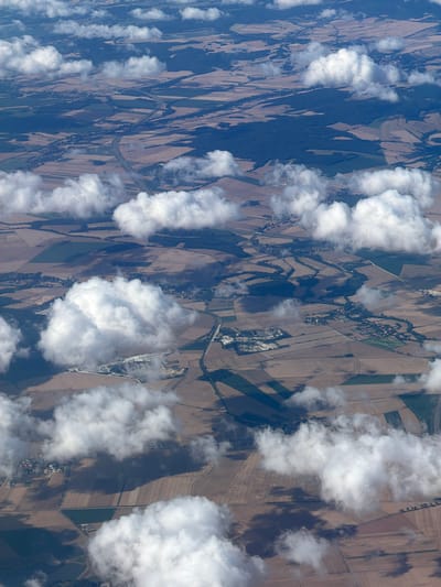 The image is an aerial view of a rural landscape, likely taken from an airplane. The scene is characterized by patchwork fields, some appearing brown and harvested, others green. Scattered throughout the landscape are small clusters of buildings, suggesting villages or hamlets. A highway is visible, cutting through the fields and connecting different areas. Cumulus clouds cast shadows on the ground, creating a dynamic interplay of light and dark across the terrain. There are no visible people or animals. The overall impression is one of a rural area undergoing agricultural activity, with human infrastructure integrated into the natural environment. The location is identified as Raczkowa, Poland.