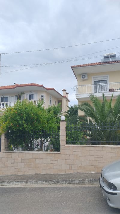 The image captures a street view in Νέα Κερασιά, Greece, under an overcast sky. In the foreground, a section of asphalt road is visible, bordered by a concrete curb and a light-colored stone wall. A silver car is parked on the right side of the frame, partially obscuring the road. Behind the wall, a black metal fence encloses a garden area. A prominent feature of the garden is a lush green tree laden with small red fruits, likely pomegranates, and several palm trees. Two residential buildings with red-tiled roofs are situated behind the garden. The building on the left is a two-story structure with balconies, white railings, and arched windows. The building on the right, also two stories high, features a balcony with white railings and an air conditioning unit mounted on its facade. The sky is filled with clouds, suggesting a potentially damp or humid day. Power lines are strung across the upper portion of the image, indicating the presence of urban infrastructure.