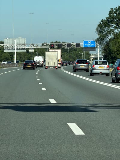 This image depicts a view from a vehicle traveling on a highway in Ede, Netherlands. The road is asphalt, marked with white lane lines, including dashed lines indicating the edge of the lane and solid white lines to the right. The sky is clear and blue, suggesting daytime with fair weather.
Several vehicles are visible on the highway, including cars and trucks, all moving in the same direction. In the foreground, a shadow of the vehicle from which the photo is taken is cast on the road.
Overhead, there are gantries with traffic lights and signage. One blue sign, which appears to be a directional sign, indicates "Maanderbroek 1600 m" and features a symbol of a crossroads. Traffic lights are also present, with one showing a red "X," indicating a closed lane.
The environment surrounding the highway is characterized by green trees and vegetation. In the distance, structures like highway signs and potentially buildings or infrastructure are visible. The overall scene suggests a typical highway environment in the Netherlands.