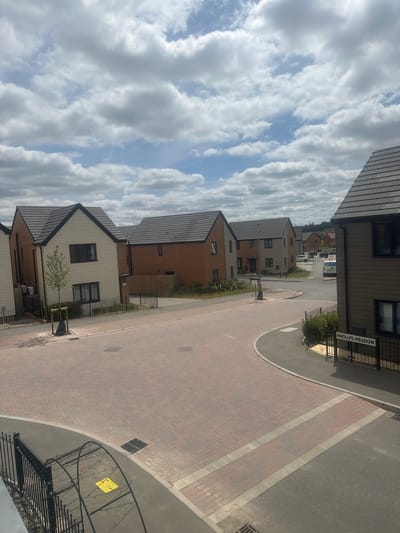 The image depicts a residential street scene in Knollys Meadow, Wellingborough, United Kingdom. The photograph is taken from an elevated perspective, looking down onto a cul-de-sac with modern houses lining the road. The houses are a mix of brick and render construction with grey tiled roofs. The road surface in the foreground is paved with red bricks, transitioning to tarmac further into the scene. A street sign indicating "KNOLLYS MEADOW" is visible on the right side of the image. Several cars, including a white van and a white car, are parked or driving on the street. The sky is partly cloudy with large white cumulus clouds. There are no people visible in the image. The environment appears to be a suburban development.