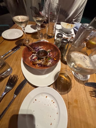 The scene depicts a wooden dining table within a restaurant or sports club located in Spanish Town, British Virgin Islands, following a meal. The table surface consists of light-colored wooden planks.
Arranged on the table from left to right:
On the far left, a stemmed wine glass contains a light-colored liquid, positioned next to another empty stemmed glass and a small, empty white side plate. Below these is a clear, empty tumbler, and a full clear water glass. Further down, a silver fork and a silver knife lie parallel on the wooden surface.
In the central area, a wooden bowl contains remnants of shredded purple cabbage, orange carrots, green leafy vegetables, and several bone fragments, consistent with consumed poultry. A small metallic ramekin filled with a dark sauce is situated within the wooden bowl. This wooden bowl rests upon a larger white plate. Below these, an empty white dinner plate displays scattered food crumbs.
On the right side of the table, an empty Heineken-branded glass stands next to two clear glass shakers with metal caps, one appearing to contain salt and the other pepper. A large, mostly empty clear glass carafe, showing residual liquid, is positioned on a round white coaster partially visible with red text reading "RESTAURANT & SPORTS CLUB." Adjacent to the carafe is a glass containing a yellow liquid and ice.
In the upper background, the upper torso of a person wearing a light-colored shirt is partially visible. Additionally, a small, empty white bowl with food residue and a fork are discernible in the background.
The overall arrangement of used plates, empty or partially full glasses, and food remnants indicates that a meal has recently concluded or is in its final stages for multiple diners.