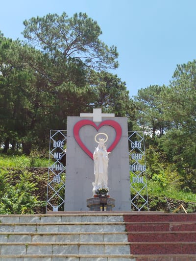 A white marble statue of the Virgin Mary holding the infant Jesus stands centrally in front of a tall, light grey monument. The monument is topped with a white cross and features a large, red heart outline behind the statue. Two diamond-shaped metal frames with Vietnamese text are positioned on either side of the statue, against the monument. The statue is adorned with a gold halo around its head. To the left of the statue, the Vietnamese text reads "ĐỨC MẸ PHÙ HỘ" and to the right, "CẦU CHÚNG CON."

The monument is situated on a terraced hillside covered in green foliage and some rocky outcrops. A set of wide stone stairs with red and grey patterned risers leads up to the monument. The scene is set against a backdrop of lush green pine trees and a clear blue sky. This appears to be an outdoor religious site, likely a shrine or a church area, in K'Rèn, Vietnam. There are no people visible in the image.