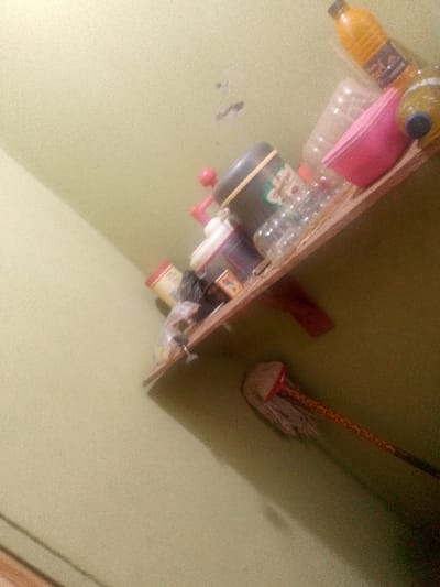 A cluttered shelf is mounted on a green wall. The shelf contains various items, including bottles, containers, and a pink bowl. A mop with a striped handle leans against the wall below the shelf. The lighting in the room appears dim, casting shadows on the wall and shelf. The image appears slightly tilted.