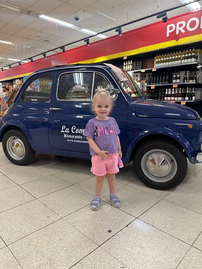 A young girl stands to the left of a dark blue vintage car parked indoors. The car has a white logo on its side that reads "La Com Ristorante" followed by a website address. The girl wears a purple t-shirt with a cat graphic and pink shorts. She has blonde hair tied up with purple ribbons and wears light purple sandals. The car and the girl are on a tiled floor. Behind the car, shelves stocked with bottles, likely wine, are visible, and above them, a red and yellow banner with the word "PRO" in large white letters. The environment appears to be a retail space, possibly a supermarket or a store with a beverage section. The specific location is identified as Berchem-Sainte-Agathe - Sint-Agatha-Berchem, Belgium.