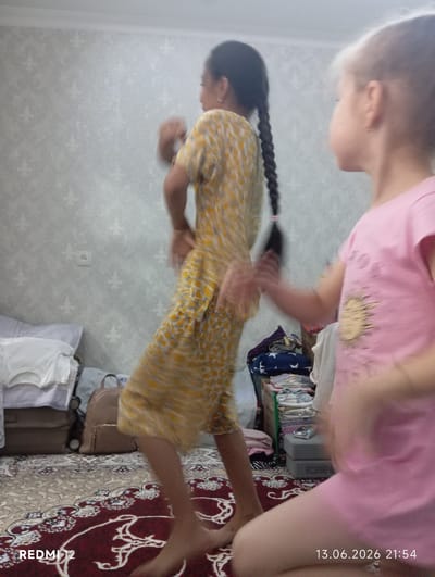 In an indoor scene with textured wallpaper and a patterned rug, two young girls are captured mid-motion, suggesting dancing or playful movement. The girl on the left, wearing a yellow and white patterned outfit, has her dark hair in a long braid and appears to be facing away from the camera. The girl on the right, partially visible and facing the camera, is wearing a pink t-shirt. The background includes stacks of folded clothing, a beige handbag, and a wall outlet. The lighting is subdued, and the image has a slightly blurry quality due to the movement. The timestamp in the lower right corner indicates the photo was taken on June 13, 2026, at 21:54, and a watermark at the bottom left shows "REDMI 12". This scene is likely in a residential setting within the city of Bekabad, Uzbekistan.