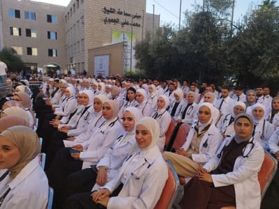 The image depicts an outdoor gathering of scores of medical students at Hebron University in Al-Khalil, Palestinian Territory. The event is set in a paved courtyard directly in front of the "Building of His Eminence Sheikh Muhammad Ali Al-Ja'bari," a multi-story, light-colored stone building with multiple dark-framed windows, prominently displaying the Arabic inscription of its name on its facade.

In the foreground and mid-ground, rows of male and female students, primarily young adults, are seated on orange-red chairs, uniformly facing forward as if attending a ceremony. The vast majority of the seated individuals are wearing white lab coats, and numerous students, both male and female, have stethoscopes draped around their necks or over their shoulders. Female students are identifiable by their hijabs, which vary in color including beige, white, light grey, and black. Male students wear white lab coats over shirts, some with ties.

To the right of the seated crowd, a dense arrangement of mature trees with dark green foliage is visible. In the distant background, a large white banner with the Hebron University emblem and Arabic text, including "جامعة الخليل" (Hebron University), is partially visible near a stage or raised platform. The scene is illuminated by natural daylight, suggesting late afternoon.